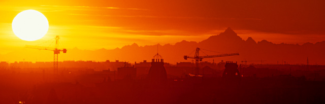 Sagome di gru spuntano tra i tetti di piazza Piemonte, sotto il sole al tramonto dietro alle cime a Sud del Monviso.  Stefano Gusmeroli